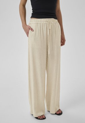 Kobieta w beżowych, szerokich spodniach w prążki z wiązaniem w talii i czarnym, bezrękawnym topie, stojąca z rękami w kieszeniach.