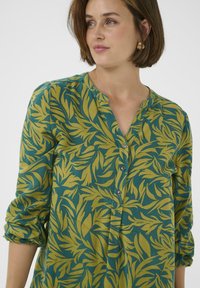 Blouse verte avec un motif de feuilles jaunes, décolleté en v et devant à boutons, manches longues retroussées. Tissu lisse avec un léger éclat et coupe décontractée.