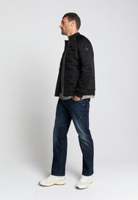 Zwarte pufferjack met gestikte textuur, staande kraag en zijzakken, gecombineerd met donkerblauwe jeans en witte sportschoenen.