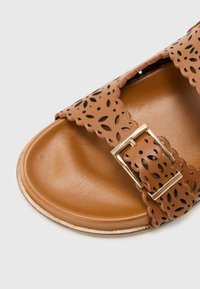 Brune skinnsandaler med perforert stropp, bølget kant og gullspenne. Glatt innersåle og flat design for enkel bruk.