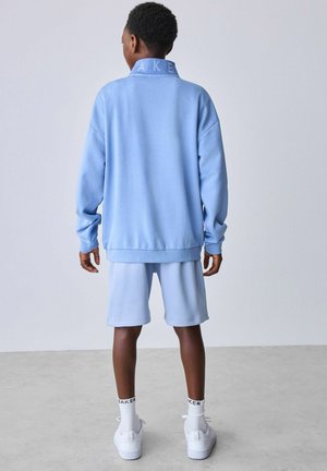 Barn iført lyseblå sweatshirt og matchende shorts, hvide sneakers og sokker, stående med ryggen mod en ensfarvet baggrund.