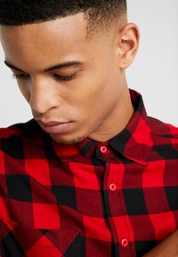 Camisa a cuadros rojos y negros con cierre de botones en el frente, que presenta un cuello y un bolsillo en el pecho. Tejido suave con acentos de botones en contraste.