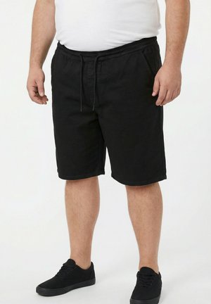 Mand iført sorte shorts med snøre, sorte sneakers og en hvid t-shirt, stående mod en ensfarvet lys baggrund.