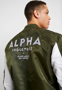 Olivgrüne Bomberjacke mit weißen Ärmeln, besticktem "ALPHA" und "INDUSTRIES" auf dem Rücken sowie zusätzlichen Textdetails darunter.