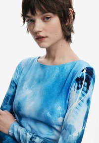 Haljina dugih rukava u raznim nijansama plave s apstraktnim uzorcima, s čamastim izrezom i pripijenim siluetom. Glatki, mekani materijal.