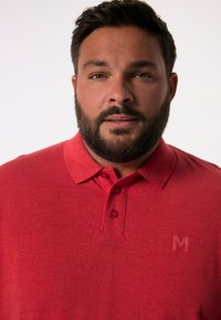 Červené polo tričko vyrobené z texturované látky s špičatým límcem a třemi knoflíky. Má decentní logo na levé straně hrudi.