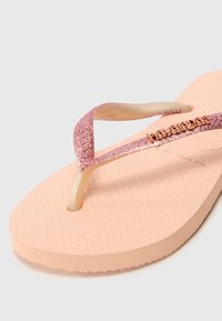 Vaaleanpunaiset varvastossut, joissa on teksturoitu kumipohja, kiiltävät vaaleanpunaisista glitter-nauhoista ja merkkilogolla varustettu sormivarvasosa.