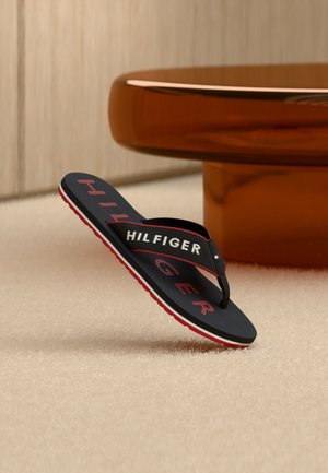 Schwarze Flip-Flop-Sandale mit roter Sohle und weiß-rotem "HILFIGER"-Logo, balancierend auf Teppich in der Nähe eines braunen Tisches positioniert.