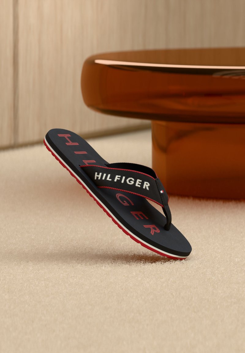 Μαύρο σαγιονάρες με κόκκινη σόλα και λευκή-κόκκινη επωνυμία "HILFIGER", ισορροπώντας σε χαλί κοντά σε καφέ τραπέζι.