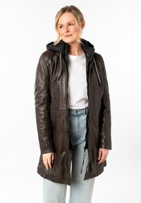 Manteau en cuir marron avec capuche noire, doté d'une fermeture éclair, de cordons de serrage réglables et d'une coupe décontractée. Porté avec un jean bleu clair.
