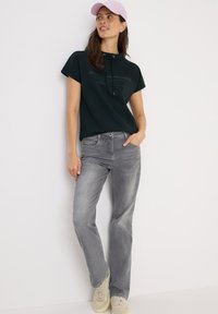 Mörkgrön kortärmad sweatshirt med huva och tryckt text, ihop med ljusgrå jeans och beige sneakers; en ljusrosa keps kompletterar looken.