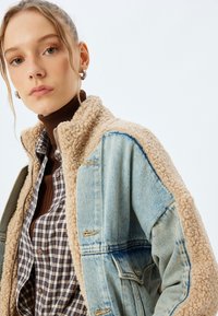 Denim- und Sherpajacke mit einem beigen strukturierten Kragen, hellblauen Denimsäulen und einem braunen karierten Hemd darunter. Silberner Reißverschluss.