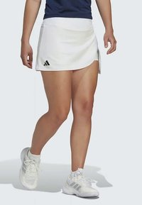 Biela športová sukňa s vysokým pásom, s bočným rozparkom a čiernym logom Adidas. V kombinácii s bielymi športovými topánkami.