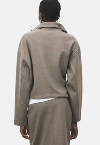 Prostorno rjavo jakno iz volnenega mešanice z širokim ovratnikom in spuščenimi ramenom, kombinirano z belo majico spodaj.