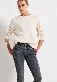 Suéter de color crema, tejido, con mangas largas y un cuello redondo, combinado con pantalones ajustados de color gris oscuro con cierre de dos botones.