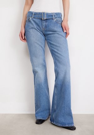Persoon die lichtblauwe jeans met hoge taille en uitlopende pijpen draagt, voorzien van een bijpassende riem en zwarte schoenen, staand tegen een witte achtergrond.
