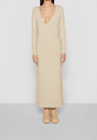 Robe maxi beige à manches longues avec un décolleté en V profond, texture tricotée et silhouette épurée, associée à des chaussures assorties à semelle épaisse.