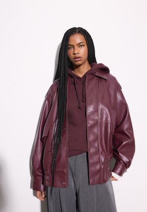 Žena s dugom pletenicom koja nosi tamnocrvenu kožnu jaknu preko odgovarajuće dukserice s kapuljačom i sive široke hlače, stoji na bijeloj podlozi.