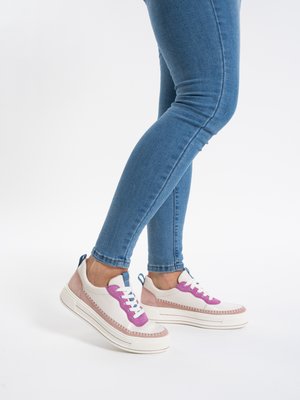 Person trägt blaue Skinny-Jeans und weiße Turnschuhe mit pinken und violetten Akzenten, steht vor weißem Hintergrund.