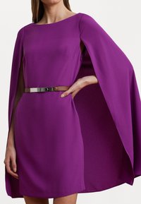 Violetti mekko viittamuotoilulla, jossa on pyöreä kaula-aukko ja lyhyet hihat, jotka on vyötetty ruskealla vyöllä sekä metallisoljella.