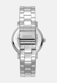 Montre ronde DKNY avec un boîtier et un bracelet en acier inoxydable. Surface lisse et brillante avec un motif de maillons. Le dos présente le logo et les détails.