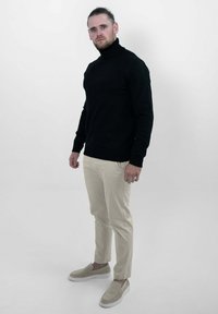 Zwarte turtleneck trui, beige slim-fit broek, beige instap schoenen, staand tegen een effen witte achtergrond. Neutrale kleurenpalet.