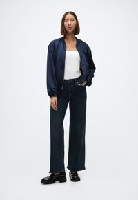 Granatowa satynowa kurtka bomberka na białej bluzce, zestawiona z ciemnoniebieskimi spodniami o szerokich nogawkach i czarnymi, masywnymi butami na platformie. Gładkie tekstury i czyste linie.