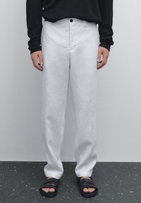 Weiße Leinenhose mit geradem Schnitt und Knopf am Bund, kombiniert mit einem schwarzen gerippten Pullover und schwarzen Sandalen. Glatte Textur.
