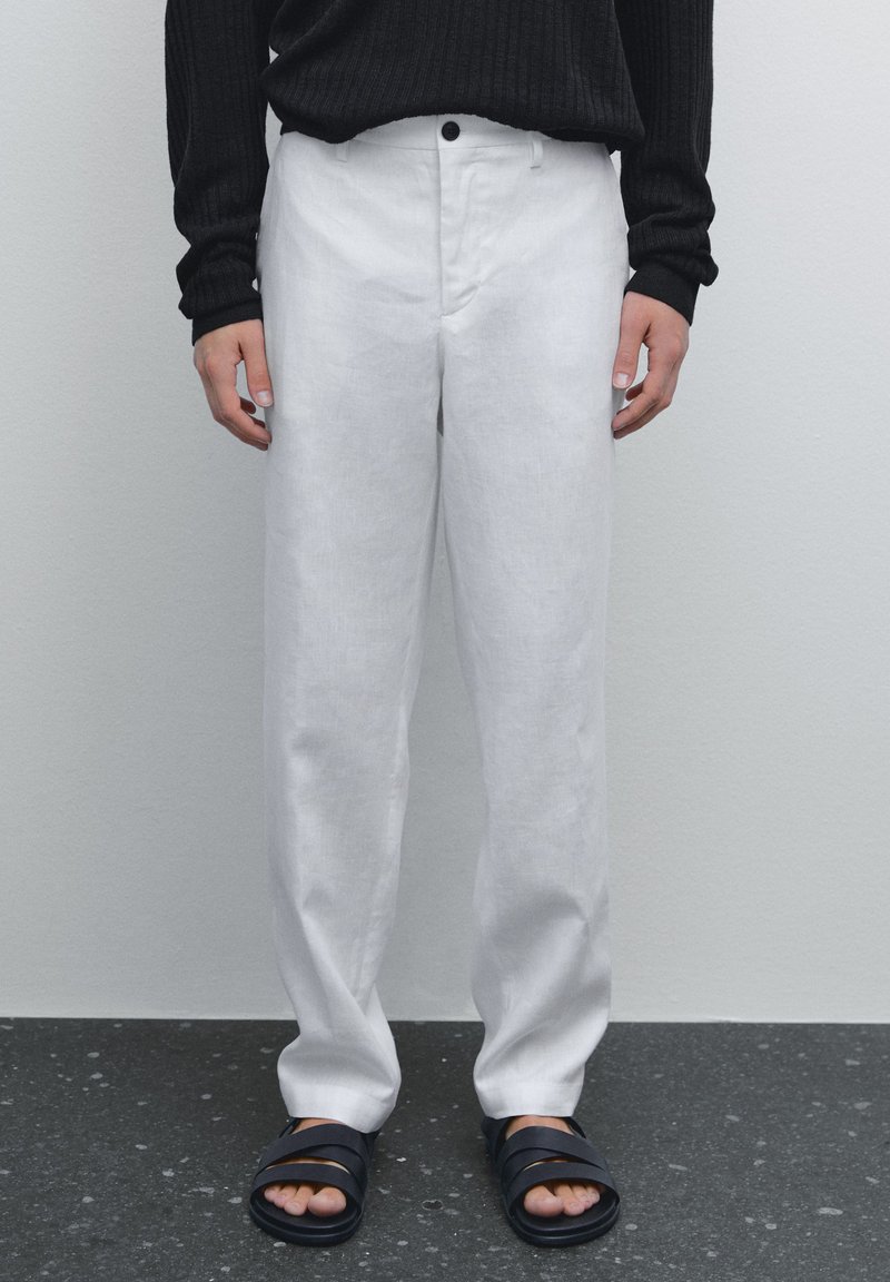 Pantalon en lin blanc à coupe droite avec un bouton à la taille, associé à un pull noir en maille côtelée et des sandales noires. Texture lisse.