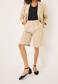 Femme portant un blazer beige et un short long assorti, un top noir, des ballerines noires à bride de cheville et un bracelet doré, debout les mains dans les poches.