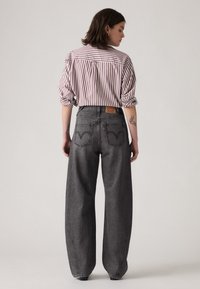 Grijze wijde jeans met een klassiek vijf-pocket ontwerp, voorzien van een leren logopatch en kenmerkende stiksels op de achterzak. Gestreept shirt eroverheen.