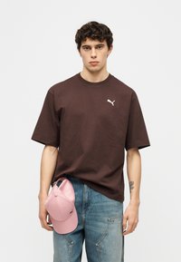 Νεαρός με σγουρά μαλλιά φοράει καφέ μπλουζάκι Puma και μπλε τζιν, κρατώντας ένα ροζ καπέλο Puma στο αριστερό του χέρι.