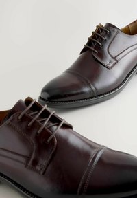 Zapatos de vestir de cuero marrón con un acabado liso, diseño con cordones y costuras detalladas en la punta y los laterales. Punta redondeada.