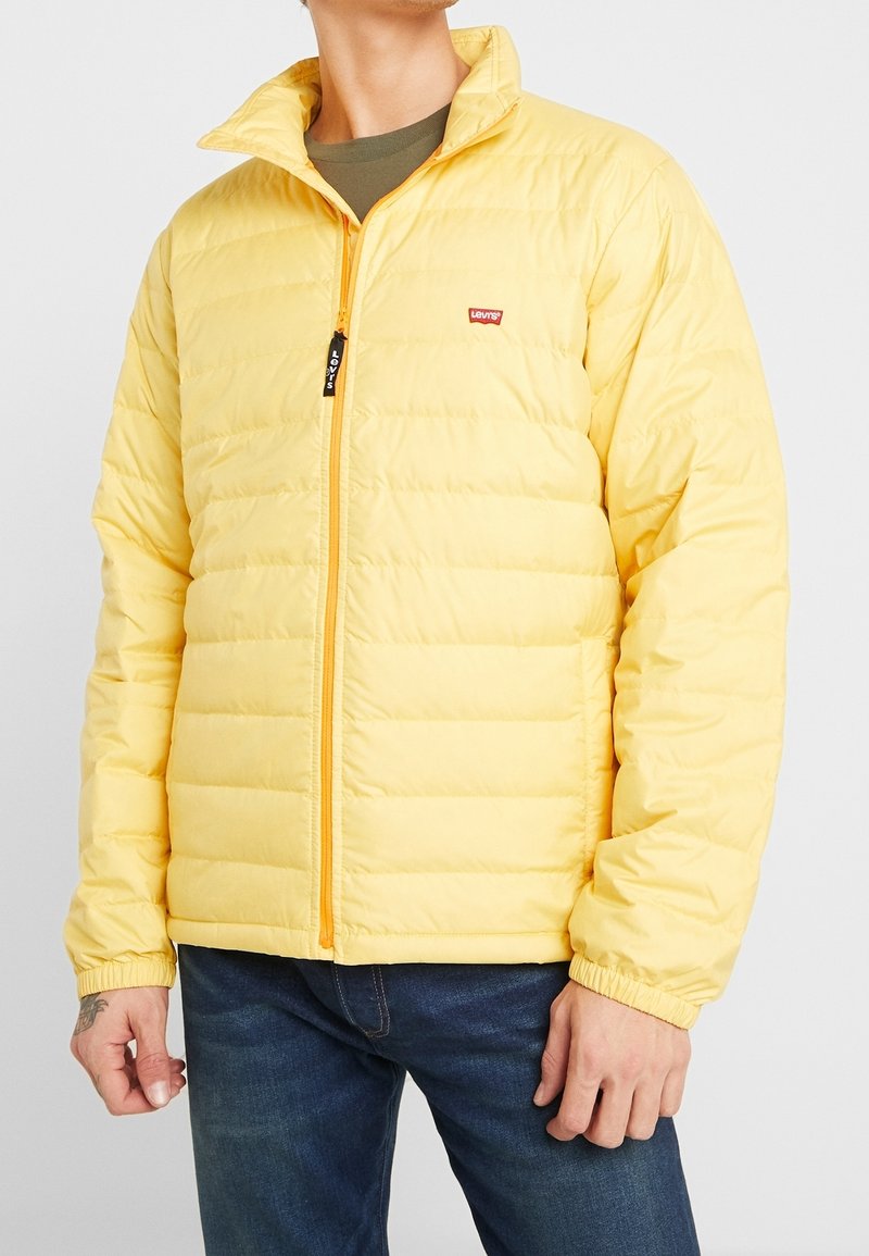 Gul pufferjakke med vandret quiltet design, orange lynlås og elastiske manchetter. Har et lille brandlogo på venstre bryst.