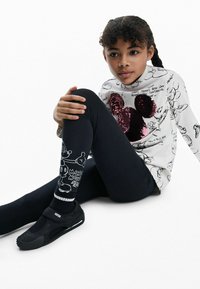 Fehér, hosszú ujjú felső fekete rajzfilmszerű ábrákkal és flitteres Mickey Mouse mintával, fekete leggingsszel, amelyeken rajzfilmszerű grafikák találhatók, valamint fekete cipővel párosítva.