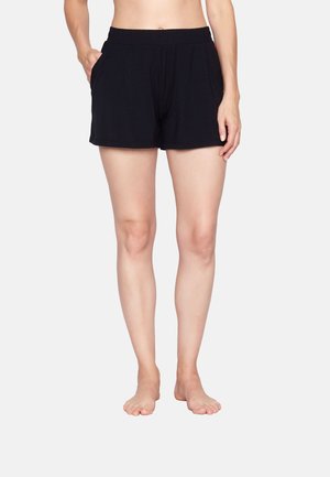 Frau trägt schwarze Shorts mit elastischem Bund und Taschen, steht barfuß vor einem schlichten, hellen Hintergrund.