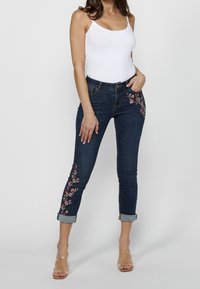Jeans de denim azul oscuro con bordado floral en los laterales, con puños enrollados y un diseño tradicional de cinco bolsillos, llevados con una blusa blanca.