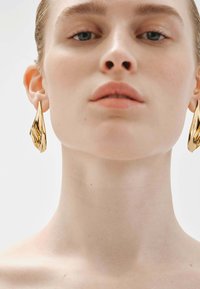 Pendientes de metal dorado con forma orgánica y un acabado texturizado, colgando de las orejas de un modelo sobre un fondo sencillo.