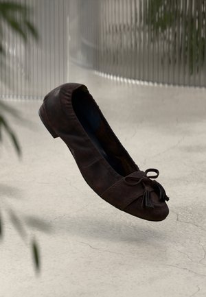 Brązowy baletowy płaski but ze skóry z zaokrąglonym noskiem, ozdobą w formie kokardki i frędzlami. Gładka tekstura i minimalistyczny design. Płaski obcas, zawieszony nad betonową powierzchnią.