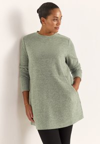 Grøn struktureret tunika med lange ærmer, rund halsudskæring og sidelommer, kombineret med sorte leggings. Glat, ribbet stofdetalje.