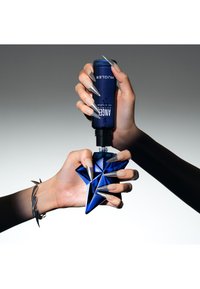 Blå glasflaske formet som en stjerne til parfume holdt i den ene hånd, med en mørkeblå klemflaske i den anden. Neglene er lange og metallisk grå.