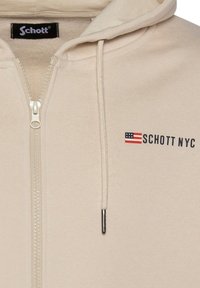 Sudadera con cremallera en color beige, fabricada en un material suave. Cuenta con capucha ajustable, cremallera y el logo "SCHOTT NYC" junto a una pequeña bandera estadounidense.