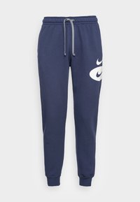 Pantaloni jogger blu navy con vita elastica e cordino grigio, caratterizzati da grandi loghi a forma di swoosh bianca sulla coscia destra.