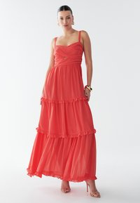 Robe longue corail avec un corsage ajusté, une jupe à volants superposés et des bretelles fines. Confectionnée dans un tissu léger, elle présente un détail en forme de goutte à la taille.