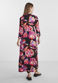 Maxiklänning i svart tyg med stora, flerfärgade blommönster. Långa ärmar och en avslappnad passform. Klack-sandaler med blockhällan på.
