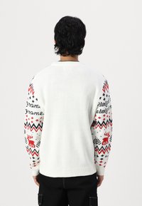 Hvid strikket sweater med mønstrede røde og sorte ærmer, der har geometriske designs og et renmotiv. Rund hals, ribbede manchetter.