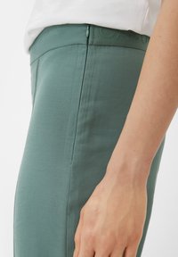Green stretch pants with a smooth texture, featuring a concealed zipper and a raised logo on the waistband. Side seam visible.