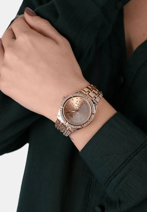 Zapestje z rožnato zlatim uramom, ki vsebuje kristalne okraske na številčnici, okvirju in povezanem zapestju, nosi nad temno zeleno majico z dolgimi rokavi.