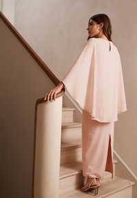 Jasnoróżowy dwuczęściowy strój z falującym topem z peleryną i długą spódnicą z rozcięciem z tyłu, uzupełniony metalicznymi sandałami na obcasie.