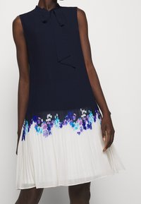 Robe sans manches avec un haut bleu marine doté d'un col à nouer, se transformant en une jupe plissée blanche ornée d'imprimés floraux bleus et violets.
